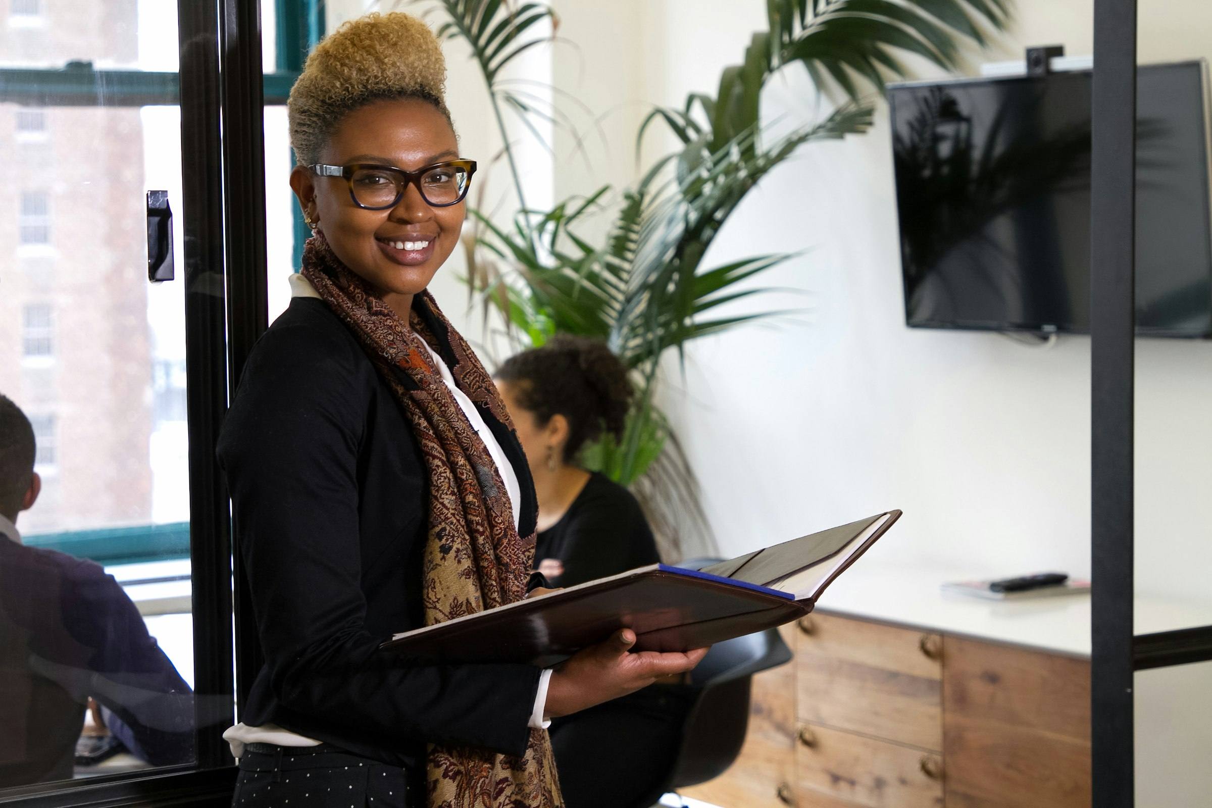 A smiling leader in a bright office, notes in hand, with her team working behind her